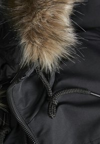 Veste noire avec une capuche en fausse fourrure texturée et amovible. Présente un cordon tissé et une fermeture éclair. La fourrure est brun clair avec des accents foncés.