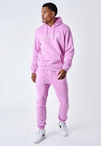 Roze hoodie en bijpassende joggingbroek van zacht materiaal. Voorzien van een voorzak en geborduurd logo op beide kledingstukken. Geaccentueerd met witte sneakers.
