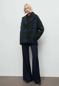 Cappotto in misto lana con motivo a quadri blu scuro e verde, silhouette oversize, ampi rever e quattro bottoni, abbinato a pantaloni svasati blu navy.