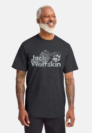 Uomo sorridente con barba bianca che indossa una maglietta nera Jack Wolfskin con disegno di montagna e impronta di zampa, mostrando le braccia tatuate.