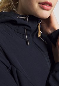 Gros plan d'une personne portant une veste à capuche sombre avec une fermeture éclair et des cordons, main touchant le cou, lèvres légèrement entrouvertes.