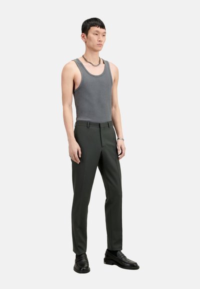 Graues Tanktop kombiniert mit dunkelgrünen Maßhosen und schwarzen Slippern. Das Model steht aufrecht mit einer Kettenhalskette und minimalen Accessoires.