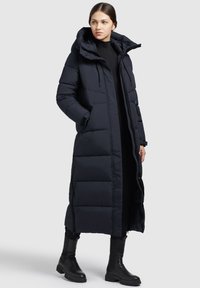 Langer, marineblauer Puffer-Mantel mit Kapuze, gesteppten Abschnitten, seitlichen Taschen und einem durchgehenden Reißverschluss. Schwarze Stiefel vervollständigen das Outfit.