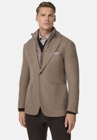 Blazer in misto lana marrone con due tasche a toppa, indossato sopra un gilet beige trapuntato. Presenta un rever e un fazzoletto da taschino a fantasia.