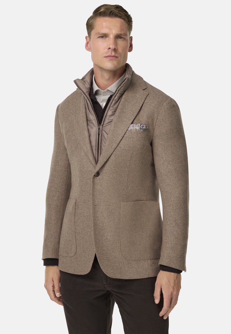 Blazer in misto lana marrone con due tasche a toppa, indossato sopra un gilet beige trapuntato. Presenta un rever e un fazzoletto da taschino a fantasia.