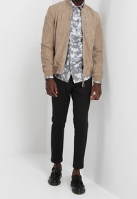 Veste bomber en suède camel, chemise à motifs gris et blanc, pantalon noir fuselé et chaussures noires avec détails à franges. Design simple et moderne.