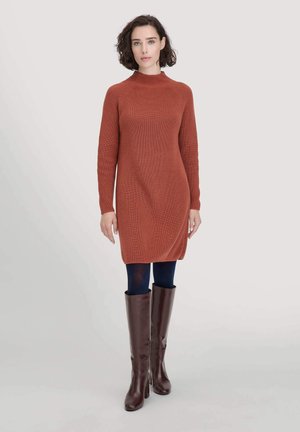 Frau mit kurzem, lockigem Haar, die ein rustfarbenes Strickkleid, dunkle Strumpfhosen und knielange braune Lederstiefel trägt, vor einem neutralen Hintergrund stehend.