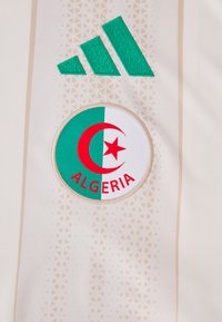 Groen Adidas-logo boven een cirkelvormig Algerijns embleem met een rode halve maan, ster en het woord "ALGERIË" op een witte stof.