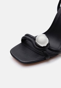 Μαύρο δερμάτινο σανδάλι με τετράγωνη μύτη, που διαθέτει λουράκι με θηλιά διακοσμημένο με μια μεγάλη ανακλαστική ασημένια σφαίρα κοντά στην περιοχή των δαχτύλων.