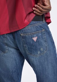 Jeans de mezclilla en un color azul claro con un acabado ligeramente texturizado. El bolsillo trasero tiene un pequeño logo triangular rojo de "Guess".