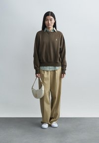 Καφετί φαρδύ πουλόβερ πάνω από πουκάμισο με πράσινες ρίγες, μπεζ παντελόνι με φαρδιά μπάρα και λευκά αθλητικά παπούτσια. Κρατάει μια λευκή καμπυλωτή τσάντα χειρός.