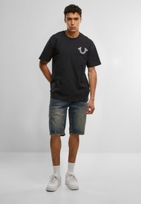 Mladý muž stojí so založenými rukami za chrbtom, oblečený v čiernom tričku s logom, denimových kraťasoch, bielych ponožkách a bielych teniskách na jednofarebnom pozadí.