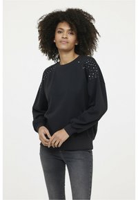 Sweatshirt noir à manches longues, avec un detailing texturé clouté sur les épaules, des poignets côtelés et une coupe décontractée, associé à un jean gris foncé.