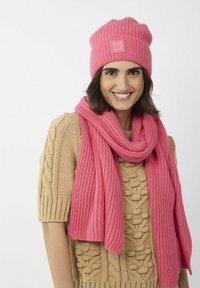 Roze gebreide beanie met een gestikt logo en bijpassende geribbelde sjaal. Trui in beige met kabelstructuur. Zachte textuur, gezellige uitstraling.