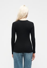 Γυναίκα με μακριά ξανθά μαλλιά, φορώντας εφαρμοστό μαύρο ριπ πουλόβερ με μακριά μανίκια και φαρδιά μπλε τζιν, στέκεται μπροστά σε λευκό απλό φόντο.