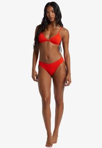 Bikini rosso con una finitura testurizzata. La parte superiore presenta un design triangolare con spalline sottili, mentre la parte inferiore a vita bassa ha una cintura liscia.