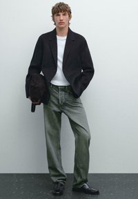 Black blazer over a white shirt, paired with faded green jeans and black loafers. The figure holds a dark brown bag, against a neutral background.