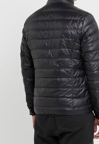 Giacca imbottita nera con finitura lucida, texture trapuntata e collo alto. Presenta un design aderente e polsini elasticizzati.