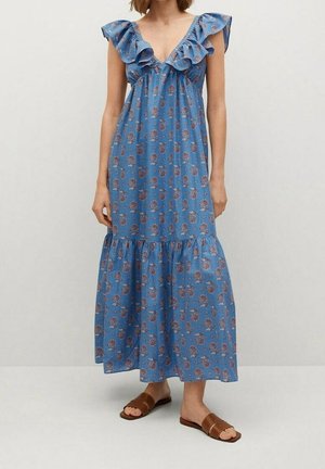 Femme portant une robe maxi sans manches bleue à fleurs avec un col volanté et des sandales plates marron, debout devant un fond uni.