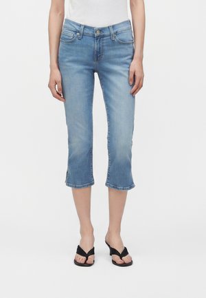 Femme portant un jean court bleu clair et des sandales à talons noires, debout devant un fond uni.