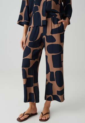 Weit geschnittene braune Hose mit großen unregelmäßigen schwarzen Formen, kombiniert mit passendem Oberteil und braunen Flip-Flops bei einer stehenden Person.