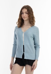 Cardigan tricotat de culoare albastru deschis, cu decolteu în V, cu butoni și model romb. Mâneci lungi cu manșete și tiv de tip rib, asortat cu pantaloni scurți negri.