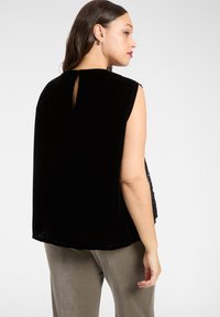 Czarny top z aksamitu bez rękawów z detalem na plecach w formie otworu, o luźnym kroju i miękkiej fakturze, zestawiony z jasnoszarymi spodniami.