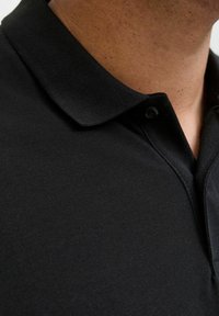 Schwarzes Polohemd aus strukturiertem Stoff, mit klassischem Kragen und einem einzelnen Knopf am Hals. Nahaufnahme hebt das Material und das Design hervor.