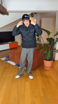 Casaco acolchoado preto, calças cinzentas, ténis brancos. Faixa preta com risca. Ambiente interior com planta verde e mobiliário laranja.