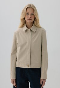 Beżowa krótka kurtka z wykładanym kołnierzem, zapięciem na guziki z przodu i kieszeniami po bokach; gładki materiał o lekko usztywnionej konstrukcji.