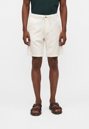 Homme portant un short écru à cordon de serrage jusqu'aux genoux et des sandales marron foncé, debout devant un fond blanc uni.