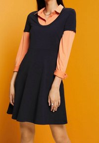Granatowa sukienka z krótkim rękawem i okrągłym dekoltem, z dołączoną pod spodem jasnopomarańczową koszulą z długim rękawem. Gładki materiał, rozkloszowana spódnica.