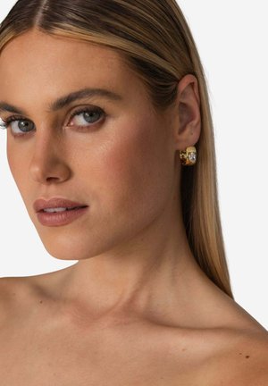 Gold hoop earring featuring a square design and a clear stone accent. Smooth texture with a shiny finish. Close-up view against a white background.