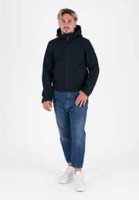 Zwarte hoodie-jack met een voorrits, gedragen over blauwe denim jeans en witte sneakers. De stof lijkt glad en heeft een aansluitend ontwerp.