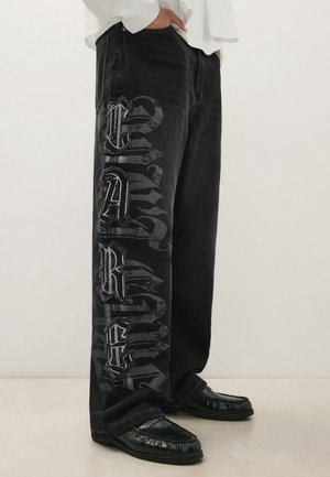 Personne portant un jean noir à jambes larges avec de grandes lettres de style gothique sur la jambe gauche et des chaussures noires sans lacets.