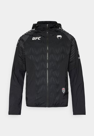 Svart vindjacka med geometriskt mönster, dragkedja framtill, huva och dragkedjefickor. Logotyper syns på bröstet och ärmarna.