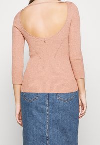 Prążkowany, różowy sweter w kolorze przygaszonego różu z niskim dekoltem na plecach, rękawami do łokcia i małym ozdobnym detalem przy zapięciu. Poniżej spódnica dżinsowa.