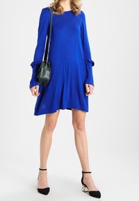 Abito blu a maniche lunghe con silhouette svasata, caratterizzato da maniche con dettagli annodati. Indossato con scarpe a punta bianco e nere e una borsa verde.