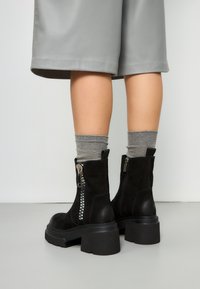 Stivaletti neri con tacco grosso, zip frontale e pelle texturizzata. Indossati con calze grigie a coste e abbinati a pantaloni corti grigi.