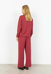 Femeie cu păr lung și blond, purtând un top larg cu mâneci lungi și pantaloni largi asortați, de culoare roz închis, stând cu fața spre un perete alb.