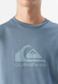 Et nærbilde av en persons nakke og skuldre som bærer en blå t-skjorte med Quiksilver-logoen i en kontrasterende design.