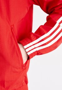 Röd jacka med en slät textur, har vita tre-stripes-accenter på ärmarna och en sidoficka. Elastiska manschetter för en snug passform.