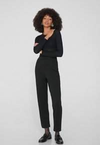 Haut noir à rayures, à manches longues, associé à un pantalon noir taille haute. Le modèle a des cheveux bouclés texturés et porte des mocassins noirs.