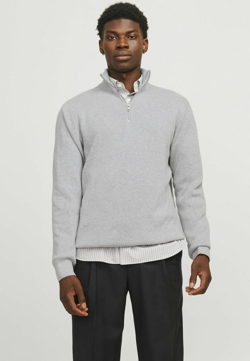 Maglione grigio chiaro con zip e lavorazione a maglia testurizzata. Presenta maniche lunghe, un colletto rialzato ed è indossato sopra una camicia a righe bianca e grigia.