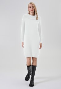 Abito bianco in maglia con scollo a barchetta, maniche lunghe e vestibilità slim, abbinato a stivali neri in pelle al ginocchio. Tessuto liscio, design minimale.