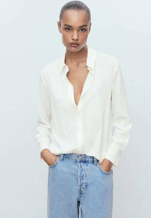 Weiße Seidenbluse mit tiefem Ausschnitt, Knopfleiste und langen Ärmeln, kombiniert mit hellblauen, hoch taillierten Jeans. Einfaches und elegantes Design.