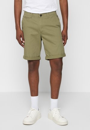 Shorts vert olive en coton, présentant une coupe droite, des ourlets roulés et un design à l'avant plat. Associés à des baskets blanches et des chaussettes.