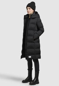 Zwarte pufferjas met een lange snit, voorzien van een capuchon, ritsluiting en twee zijzakken, gedragen over zwarte leggings en enkellaarsjes.