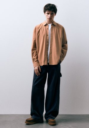 Koszula w kratę w odcieniu beżu noszona na białej koszulce, połączona z luźnymi ciemnymi spodniami jeansowymi i brązowymi butami na platformie. Gładkie tekstury i swobodny krój.