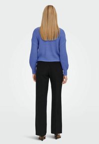 Mujer con cabello largo rubio, vistiendo un suéter de punto azul, pantalones negros de pierna recta y sandalias de tacón negras, de pie, de espaldas.